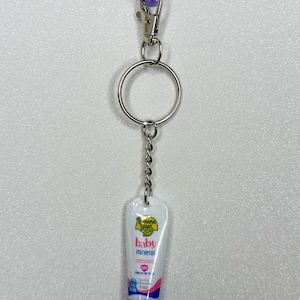 May include: A keychain featuring a miniature tube of Banana Boat Baby Mineral sunscreen. The white tube has pink and blue accents, with the brand name and product details printed on it. The keychain includes a silver ring, a chain, and a purple clasp.