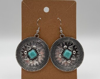 Puede incluir: Un par de pendientes colgantes circulares de tono plateado con un intrincado patrón de rayos en relieve y una piedra cuadrada de color turquesa en el centro. Los pendientes cuelgan de ganchos de metal plateado sobre una tarjeta.