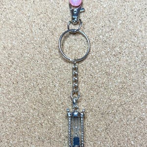May include: A silver-coloured keychain featuring a pink, glittery ball clasp, a chain, and a miniature hourglass charm. The hourglass is clear with blue sand inside. The keychain is set against a textured, tan background.