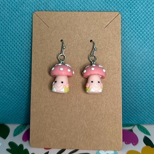 May include: A pair of mushroom-shaped earrings. Each earring features a pink cap with white polka dots, a smiling face, and a white and green base. The earrings are attached to silver-colored fish hook ear wires and displayed on a brown card.