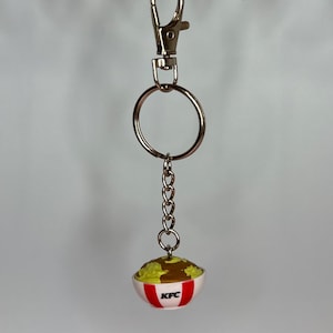 May include: A silver keychain with a lobster clasp and a small chain holding a miniature KFC bowl. The bowl is white with red stripes and the KFC logo. The bowl contains a miniature serving of mashed potatoes and gravy.