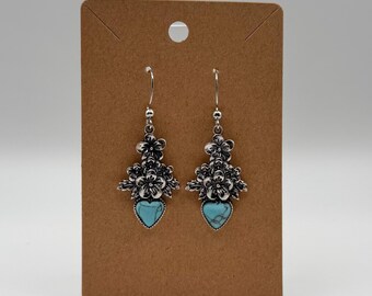 Puede incluir: Un par de pendientes colgantes de tono plateado con un motivo floral en la parte superior y una piedra en forma de corazón de color turquesa en la parte inferior. Los pendientes están sujetos a una tarjeta de cartón marrón rectangular.