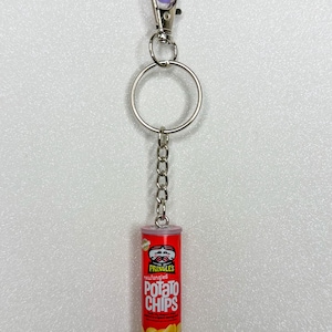 May include: A miniature Pringles potato chips can keychain. The red can features the Pringles logo and the words "Potato Chips." The keychain has a silver chain, a silver ring, and a purple clasp.