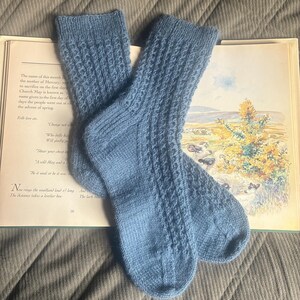 May include: Two light blue knitted socks with a textured design, displayed on an open book with illustrations and text. The socks appear to be made from a soft yarn. The book's pages are visible, creating a vintage feel.