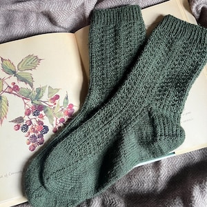 Pode incluir: Um par de meias de malha verde floresta com um padrão texturizado, repousando sobre um livro aberto com ilustrações botânicas. As meias são feitas de um material macio e quente.