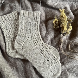 Op de afbeelding: Een paar lichtgrijze gebreide sokken met geribbelde en patroonsecties, hangend aan een metalen hanger. De sokken worden gepresenteerd op een textiel, grijze stof, met een kleine bundel gedroogde gele bloemen en touw op de achtergrond.