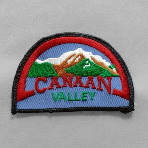 May include: Embroidered patch featuring a mountain landscape with the text "CANAAN VALLEY". The patch has a blue background with red, green, brown, and white details. The patch is in a half-circle shape with a black border.