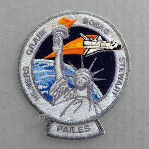 May include: A white, gray, and blue embroidered patch with a space shuttle flying over a stylized Statue of Liberty. The text "GRISSOM & STEWART" is on the left side of the patch, and "BOBKO & PAILES" is on the bottom. The text "STS-8" is on the right side of the patch.
