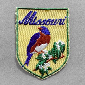 May include: Embroidered patch featuring the word "Missouri" in blue script above a blue, brown, and white bird perched on a flowering branch. The patch has a yellow background and a green border.