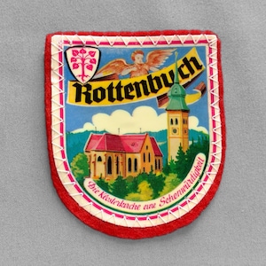 May include: A shield-shaped felt patch with a red border and white zigzag stitching. The center features a colorful illustration of a church with a red roof and a green steeple, the text Rottenbuch, an angel, and a crest with a pink tree.