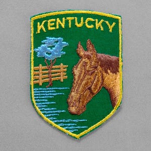 May include: Embroidered patch featuring the word "KENTUCKY" in yellow on a green shield. The patch depicts a horse's head, a tree, a fence, and water lines. The patch has a yellow border.