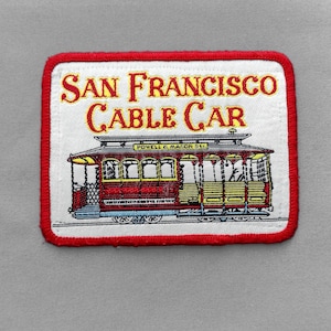 May include: A rectangular embroidered patch featuring a red San Francisco cable car on a white background. The text SAN FRANCISCO CABLE CAR is stitched in red above the vehicle. The patch has a thick red border and measures approximately 3 inches.