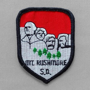 May include: Embroidered patch featuring Mount Rushmore with a red and white background. The patch has a black border and includes the text "MT RUSHMORE S.D." below the monument. The design includes green trees.