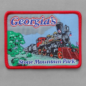 May include: Embroidered patch featuring a detailed illustration of a vintage steam train. The patch has a red border and includes the text "Georgia's Stone Mountain Park" in blue and white lettering. The train is depicted in red, black, and gold, set against a blue sky and green foliage.