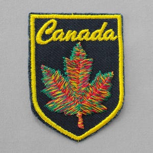 May include: Embroidered patch featuring the word "Canada" in yellow script above a colorful maple leaf. The patch has a navy blue background with a yellow border.