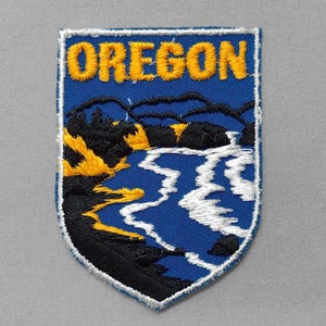 May include: Embroidered patch featuring the word "OREGON" in yellow thread above a scenic landscape. The design includes a blue river, black and yellow trees, and a white border. The patch is shield-shaped.