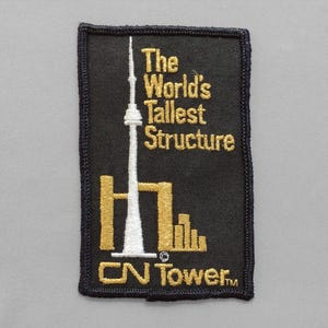 May include: Black rectangular patch with gold and white embroidered text and imagery. The text reads "The World's Tallest Structure" above an image of the CN Tower. The bottom of the patch reads "CN Tower™" in gold.