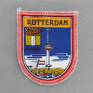 May include: A fabric patch featuring the word "ROTTERDAM" in yellow, a blue shield with a tower, and the word "EUROMAST". The patch has a red border with white stitching and a Dutch flag.