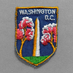 May include: Embroidered patch featuring a blue shield with the text "WASHINGTON D.C." and a depiction of the Washington Monument. Pink and white flowering trees flank the monument, with a green base.