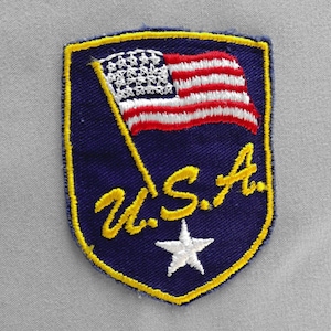 May include: Embroidered patch featuring a shield shape with a yellow border. The patch displays a waving American flag in red, white, and blue, with the text "U.S.A." in yellow and a white star.
