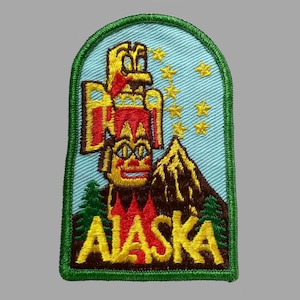 May include: Embroidered Alaska patch featuring a totem pole, mountain, and the word "ALASKA" in yellow. The patch has a light blue background with yellow stars and a green border. The totem pole is red and yellow.