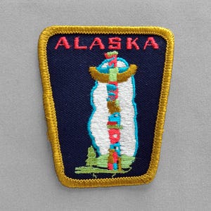 May include: Embroidered patch featuring the word "ALASKA" in red above a stylized totem pole design. The patch has a navy blue background with a gold border and a white, blue, green, and red totem pole design.