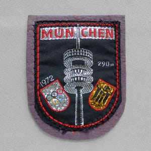 May include: A vintage patch with the word "MUNCHEN" in red letters. The patch features a silver tower, the text "290m", and two emblems, one with Olympic rings and the other with a gold design. The year "1972" is also visible.