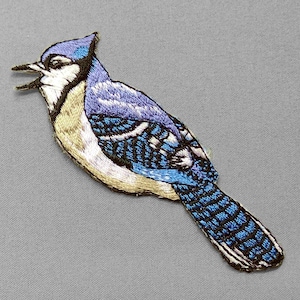 May include: Embroidered patch of a blue jay bird with open beak. The bird features shades of blue, white, and tan thread. The patch is likely for clothing or accessories.