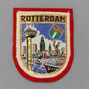 May include: Embroidered patch featuring the word "ROTTERDAM" above a colorful depiction of the city's skyline, including a tower and ships. The patch has a red border with gold and white accents.