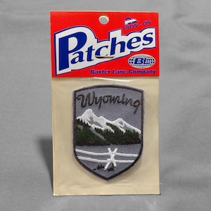 May include: Iron-on patch featuring a gray shield with the word "Wyoming" and a white mountain range with a green forest in front of it. The patch is on a white card with the text "Patches Baxter Lane Company" and "Iron-on" printed on it.