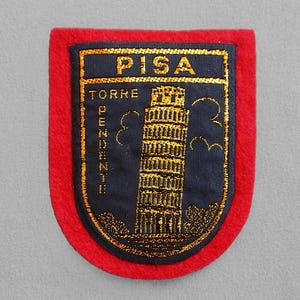 May include: Embroidered patch featuring the Leaning Tower of Pisa. The patch has a navy blue background with gold lettering that reads "PISA" and "TORRE PENDENTE". The tower is also depicted in gold. The patch is framed by a red border.