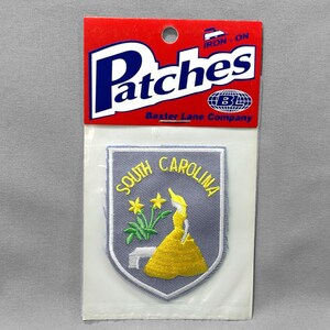 May include: An iron-on patch featuring the South Carolina state flag design. The patch is a shield shape with a gray background, yellow text, and a yellow dress design. The patch is packaged on a red and white backing.