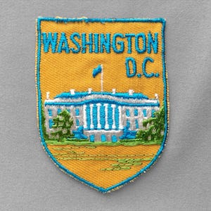 May include: A shield-shaped embroidered patch with a golden yellow background and a blue border. It features the text WASHINGTON D.C. in blue, above a white building with blue accents and green foliage at the base, all on a gray fabric surface.