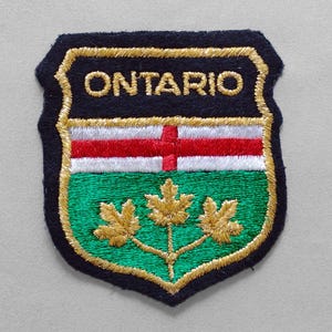 May include: Embroidered patch featuring the word "ONTARIO" in gold lettering above a design with red and white stripes and a green field with three gold maple leaves. The patch has a black border with gold trim.