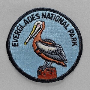 May include: Embroidered patch featuring a pelican perched on a wooden post. The patch has a light blue background with the words "EVERGLADES NATIONAL PARK" in a circular design. The pelican is depicted with orange, white, and gray feathers.