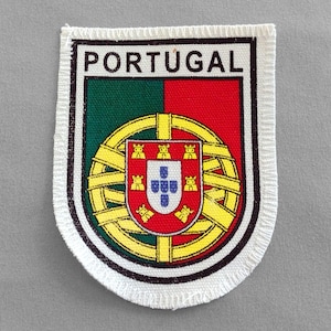 May include: A white fabric patch with a black border and the word "PORTUGAL" printed in black at the top. The patch features the Portuguese coat of arms with a red and green background.