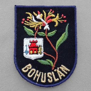 May include: A blue and gold embroidered patch with a red, yellow, and white flower and a small white building with a red roof. The text "BOHUSLAN" is embroidered in gold on the bottom of the patch.