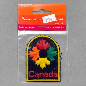 May include: A Canada patch featuring a black background with a yellow border. The patch has five colorful maple leaves in red, orange, yellow, and green. The word "Canada" is embroidered in yellow. The patch is in a clear plastic bag with a red and orange header.