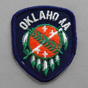 May include: A blue embroidered patch with the word "OKLAHOMA" in white lettering and a stylized image of the state flag. The flag features a white cross on a red background with a green feather design.