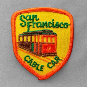 May include: A shield-shaped embroidered patch with a bright yellow background and a vibrant orange border. It features a brown and red cable car illustration with the green embroidered text San Francisco above and Cable Car below the vehicle.