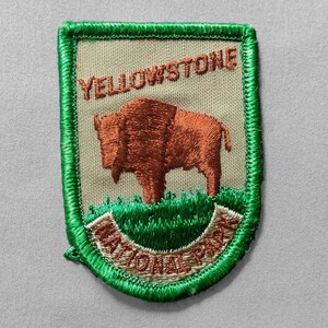 May include: Embroidered patch featuring a brown bison on green grass, with the text "YELLOWSTONE NATIONAL PARK" in brown and red. The patch has a green border and a tan background.