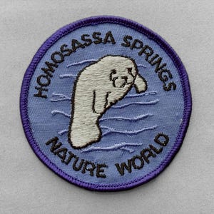 May include: A circular embroidered patch with a light blue background and a dark purple border. The center features a white manatee swimming in stylized blue water. The text HOMOSASSA SPRINGS NATURE WORLD is stitched in dark brown capital letters.
