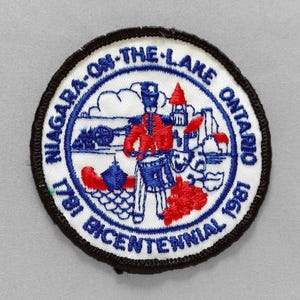 May include: Embroidered patch with a black border and a white center. The patch features a blue and red design with text that reads "Niagara-on-the-Lake Ontario 1781 Bicentennial 1981". The design includes a soldier, buildings, and a cannon.