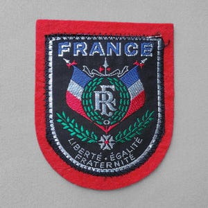 May include: Embroidered patch with a red border and a black center. The patch features the word "FRANCE" in blue, two flags, a wreath, and the words "LIBERTE, EGALITE, FRATERNITE".
