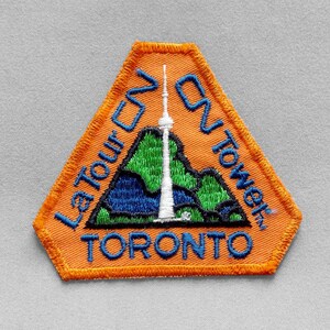 May include: Embroidered orange patch featuring the CN Tower in Toronto. The patch has a mountain scene with the tower in white, green, and blue. The text "La Tour CN Tower" and "Toronto" are in blue.