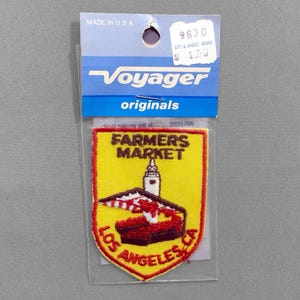 May include: A yellow, red, and white embroidered patch with the text "FARMERS MARKET LOS ANGELES CA". The patch features a building with a tower and a red and white awning. The patch is attached to a blue and white Voyager originals card.