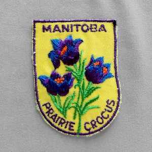 May include: Embroidered patch featuring a yellow shield with the word "MANITOBA" at the top. The patch displays three purple and blue crocus flowers with green stems. The words "PRAIRIE CROCUS" are arched at the bottom.