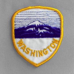 May include: Embroidered patch featuring a shield design with a mountain scene in blue and white. The word "WASHINGTON" is embroidered in yellow along the bottom edge, with a gold border.