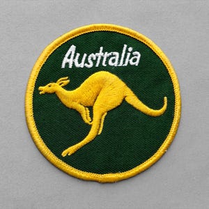 May include: A circular embroidered patch featuring a dark green background with a yellow kangaroo silhouette in the center. The word Australia is stitched in white letters above the kangaroo, and the entire patch is bordered by a yellow edge.