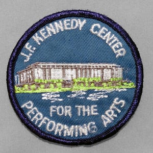 May include: A circular embroidered patch with a blue background and a dark blue border. It features a white building illustration, green landscaping, and white text that reads J.F. KENNEDY CENTER FOR THE PERFORMING ARTS in a circular arrangement.